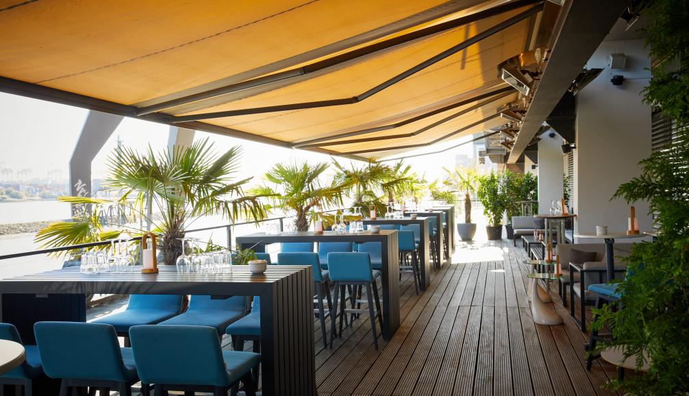 am kai Hamburg Terrasse – stilvoll eingedeckte Tische mit blauen Stühlen, Palmen und Blick auf die Elbe