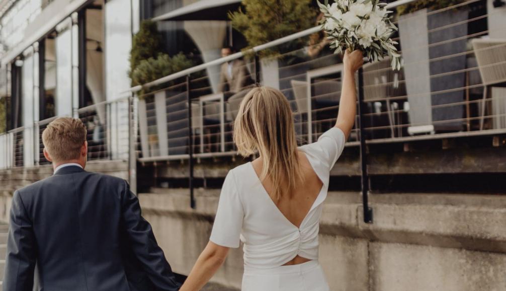 Hochzeit am kai Hamburg – Brautpaar vor der Terrasse des Restaurants mit Blumenstrauß und Elbblick